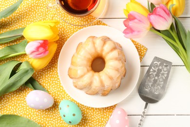 Photo of Delicious bundt cake, Easter eggs, tea and tulips on white wooden table, flat lay