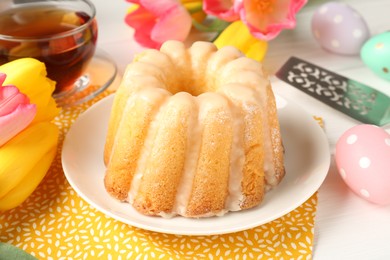 Photo of Delicious bundt cake, Easter eggs, tea and tulips on white wooden table, closeup