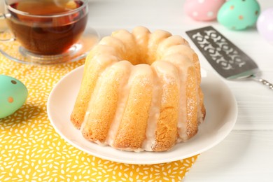 Photo of Delicious bundt cake, Easter eggs, tea and server on white wooden table, closeup