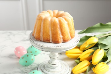 Photo of Delicious bundt cake, Easter eggs and tulips on white marble table, closeup