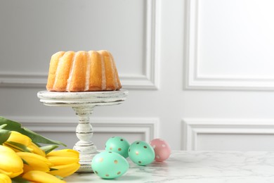 Photo of Delicious bundt cake, Easter eggs and tulips on white marble table, space for text