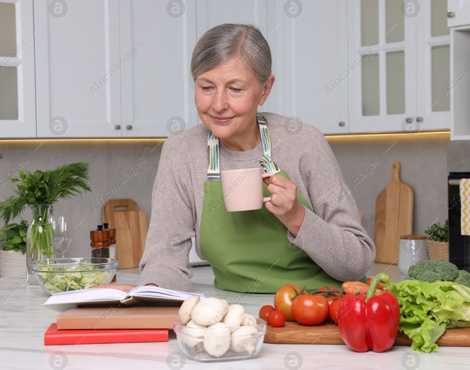 Photo of Happy woman with cup of tea and recipe book at table in kitchen