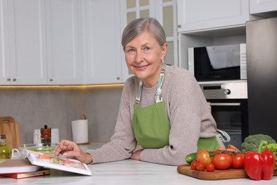 Photo of Happy woman with recipe book at table in kitchen