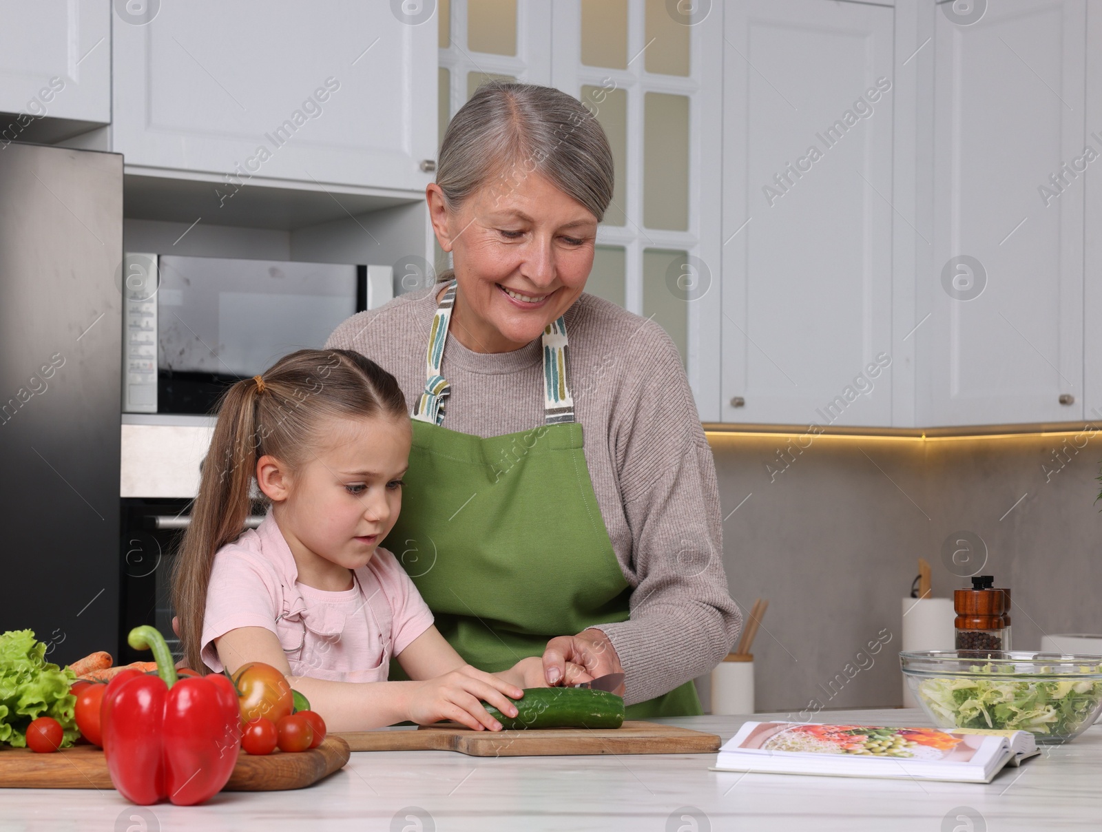 Photo of Cute little girl with her granny cooking by recipe book in kitchen