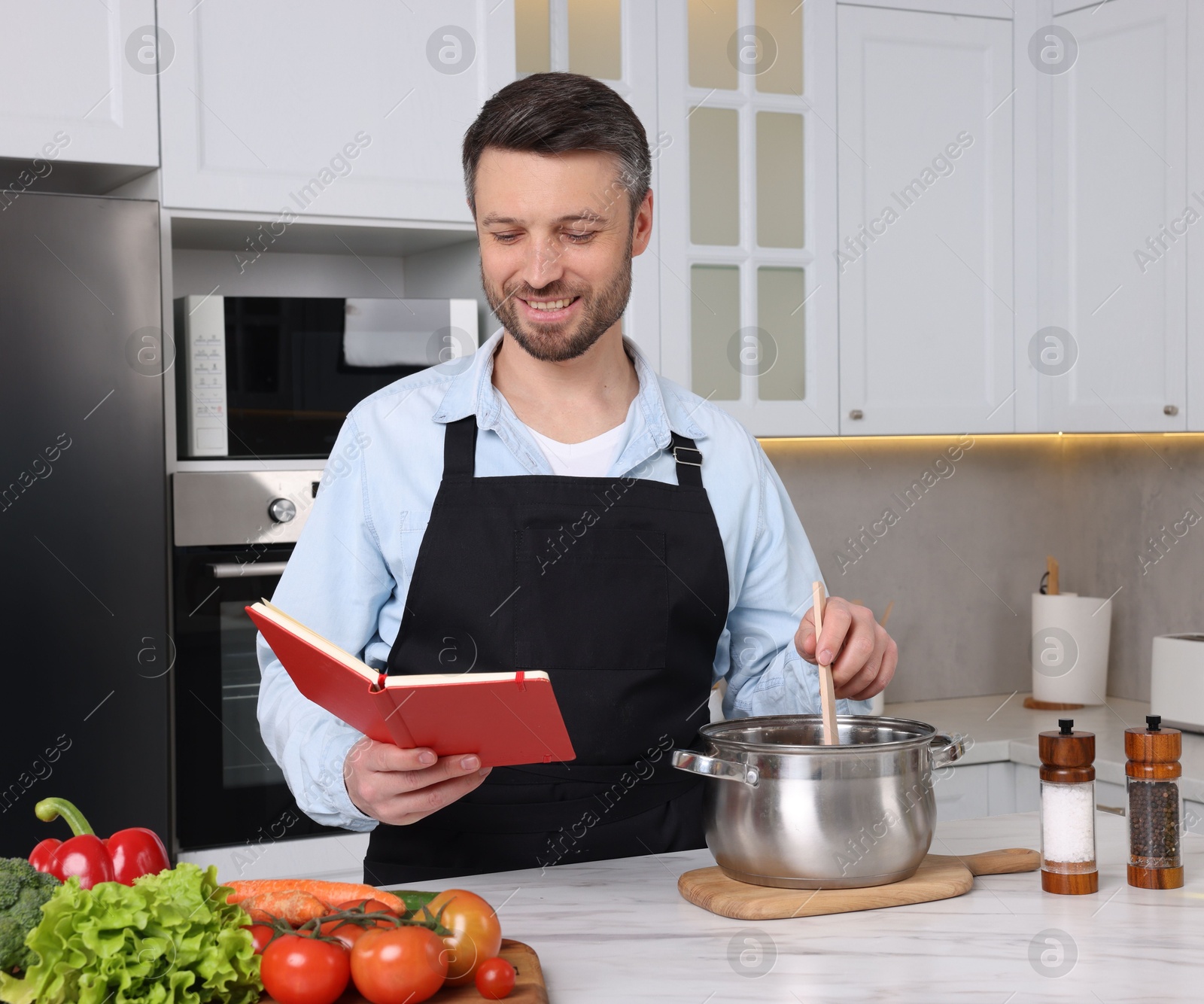 Photo of Happy man cooking by recipe book in kitchen