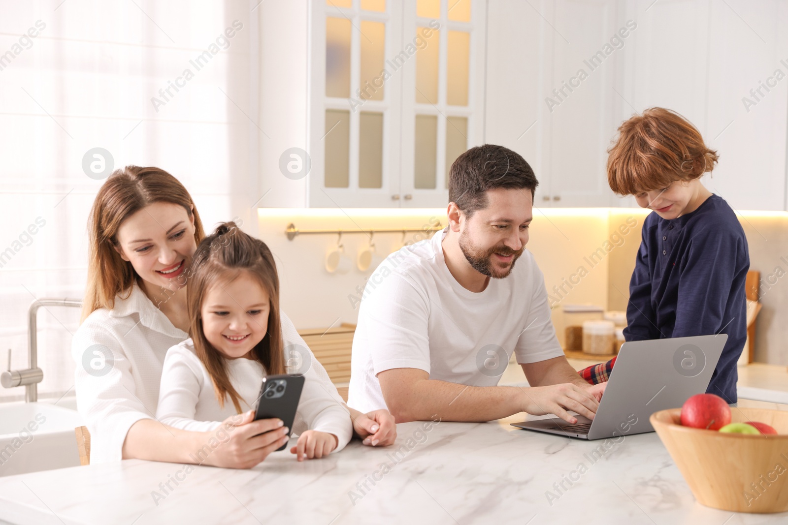 Photo of Happy family wearing stylish pajamas looking at devices at white marble table in kitchen