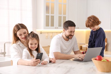 Happy family wearing stylish pajamas looking at devices at white marble table in kitchen