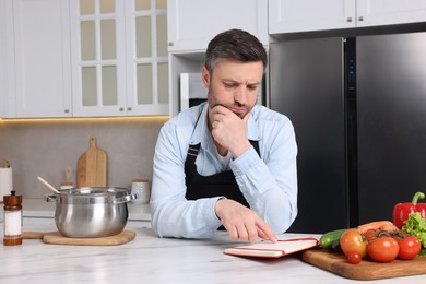 Adult man reading recipe book at white marble table in kitchen