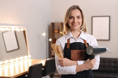 Smiling hairdresser with dryer and combs in beauty salon, space for text