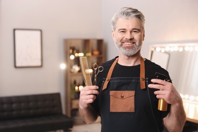 Photo of Smiling hairdresser with comb, scissors and spray bottle in beauty salon, space for text