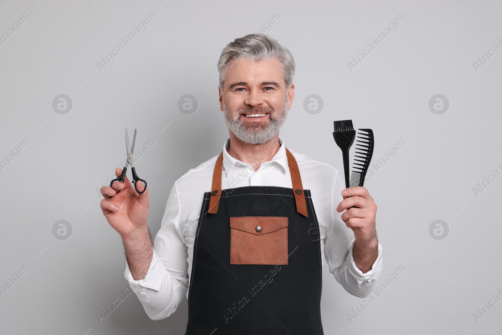 Photo of Smiling hairdresser with scissors, comb and hair dye brush on gray background