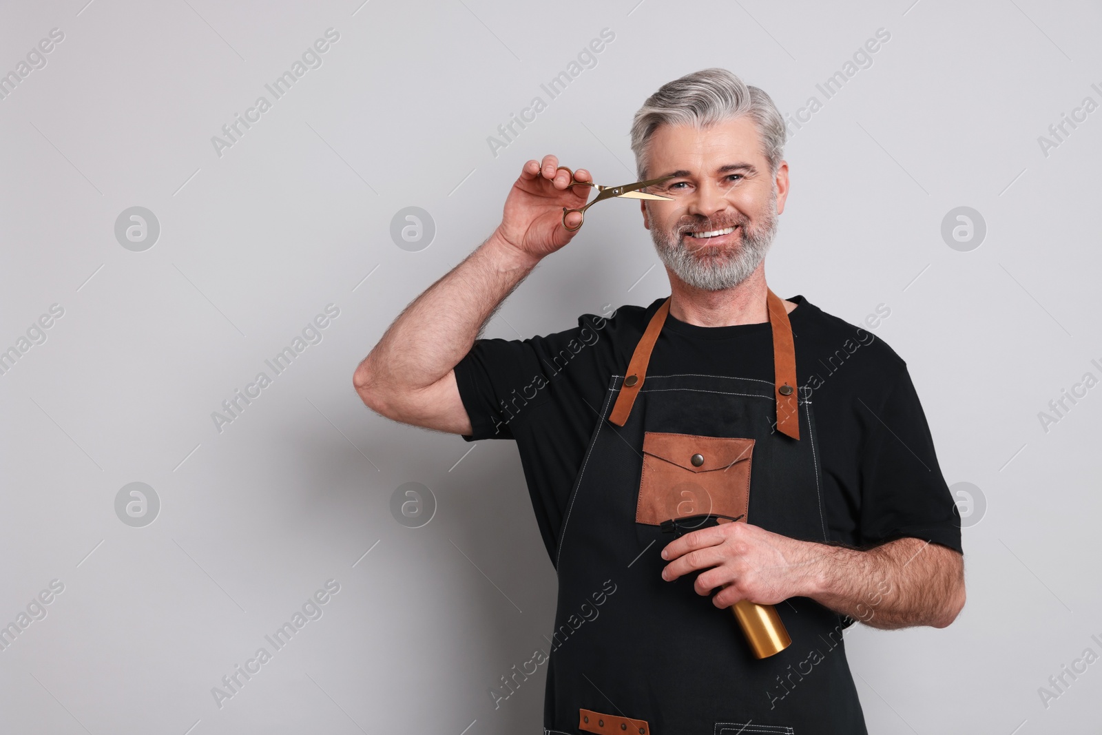 Photo of Smiling hairdresser with scissors and spray bottle on gray background, space for text