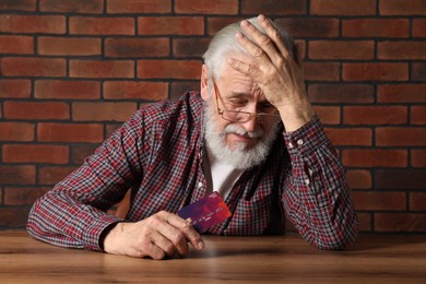 Upset senior man with credit card at wooden table. Financial problems