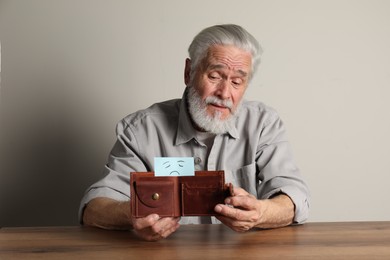 Photo of Upset senior man holding wallet with drawn sad face at wooden table. Financial problems