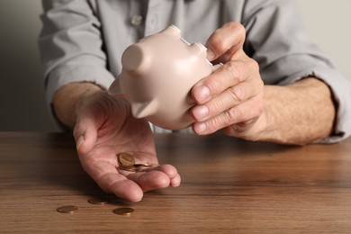 Senior man trying to get something out from piggy bank at wooden table, closeup. Financial problems