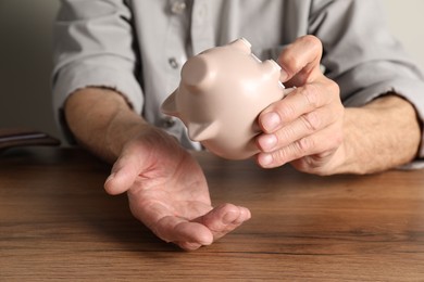 Senior man trying to get something out from piggy bank at wooden table, closeup. Financial problems