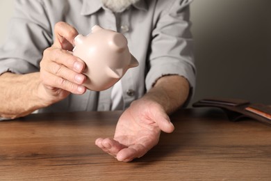 Photo of Senior man trying to get something out from piggy bank at wooden table, closeup. Financial problems