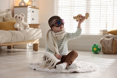 Photo of Happy little boy in scarf and aviator goggles playing with toy plane at home