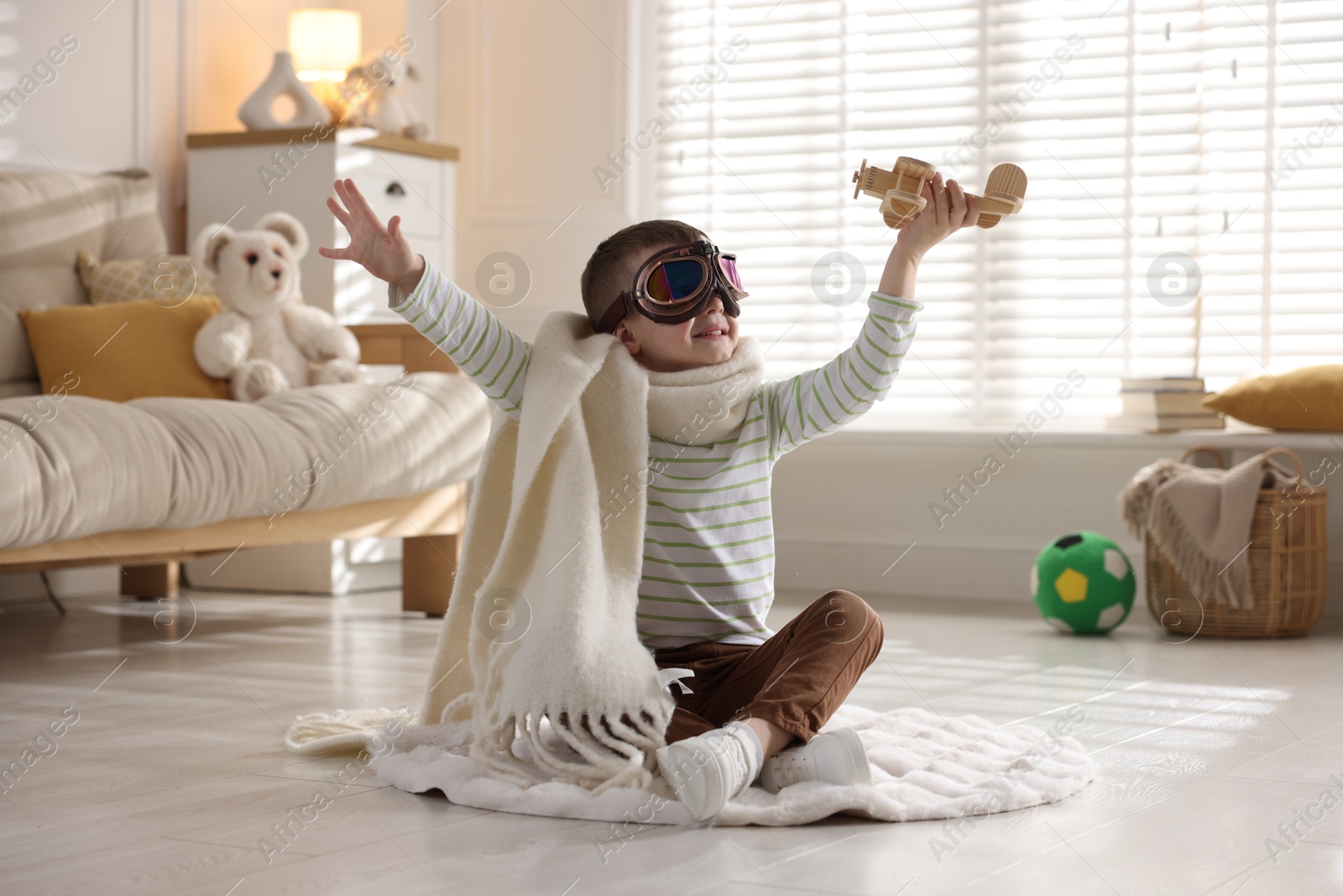 Photo of Happy little boy in scarf and aviator goggles playing with toy plane at home