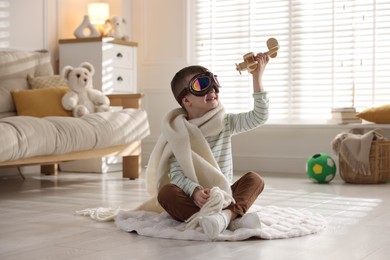 Photo of Happy little boy in scarf and aviator goggles playing with toy plane at home