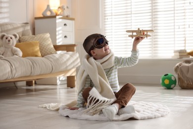 Happy little boy in scarf and aviator goggles playing with toy plane at home