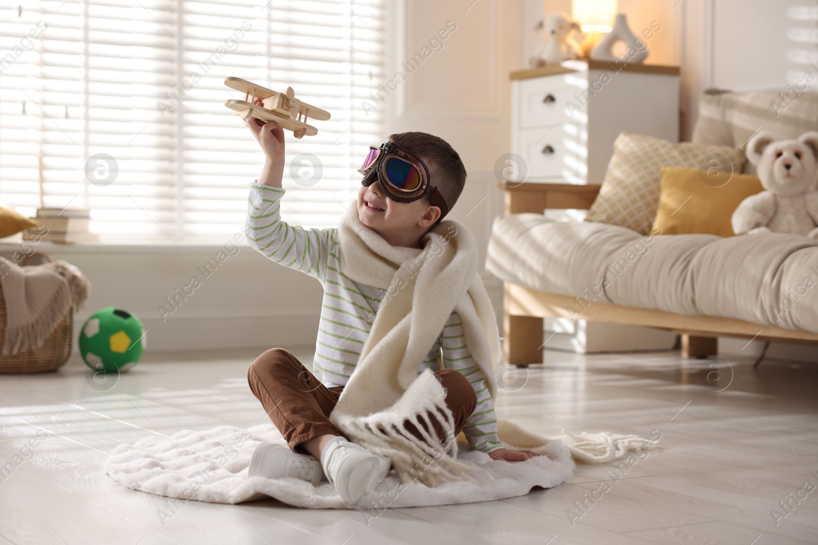 Photo of Happy little boy in scarf and aviator goggles playing with toy plane at home