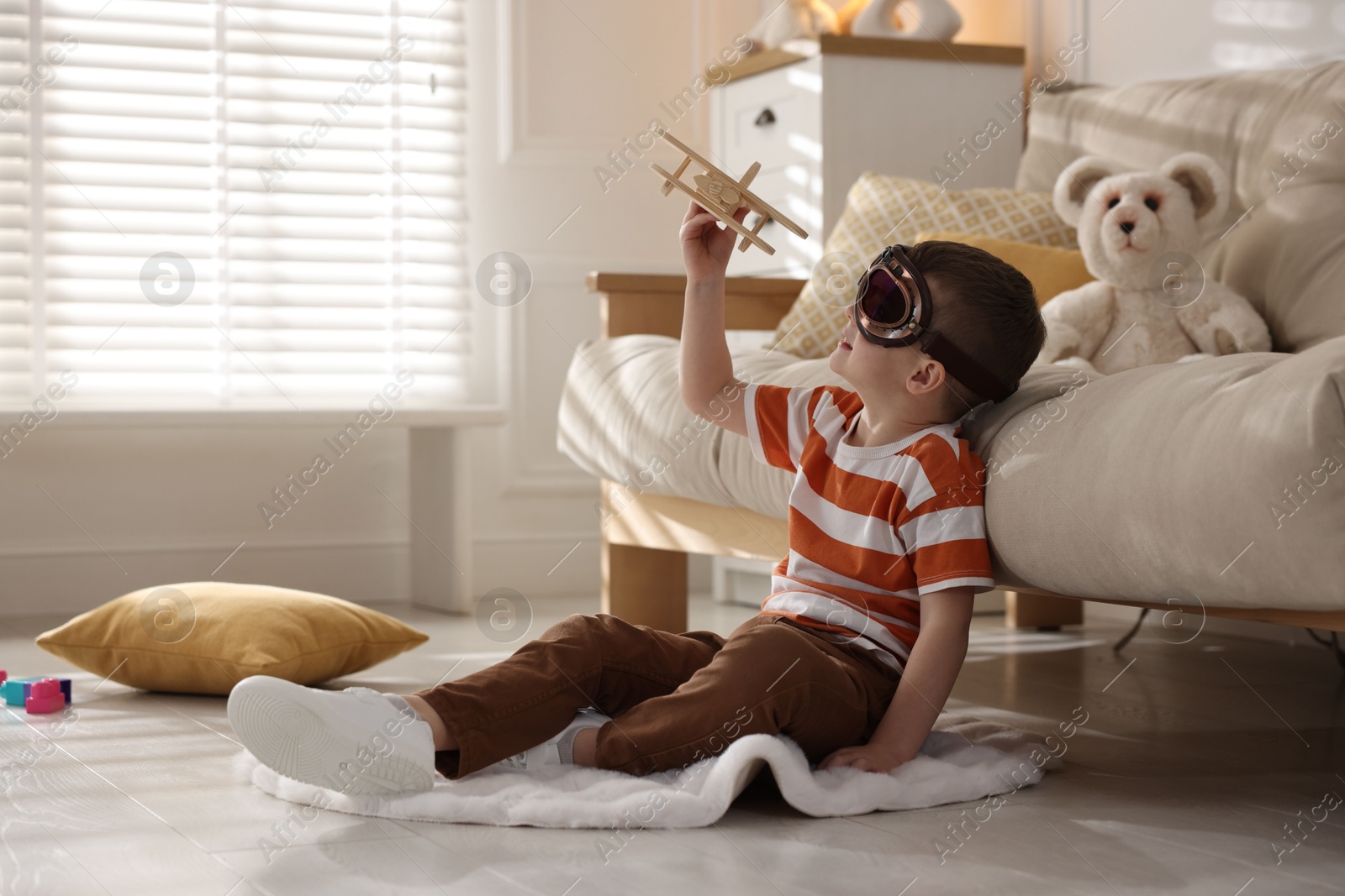 Photo of Cute little boy in aviator goggles playing with toy plane at home