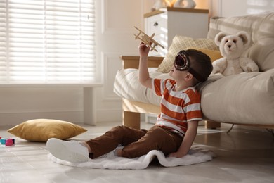 Cute little boy in aviator goggles playing with toy plane at home