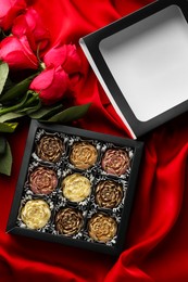 Photo of Delicious flower shaped chocolate bonbons in box and roses on red fabric background, top view