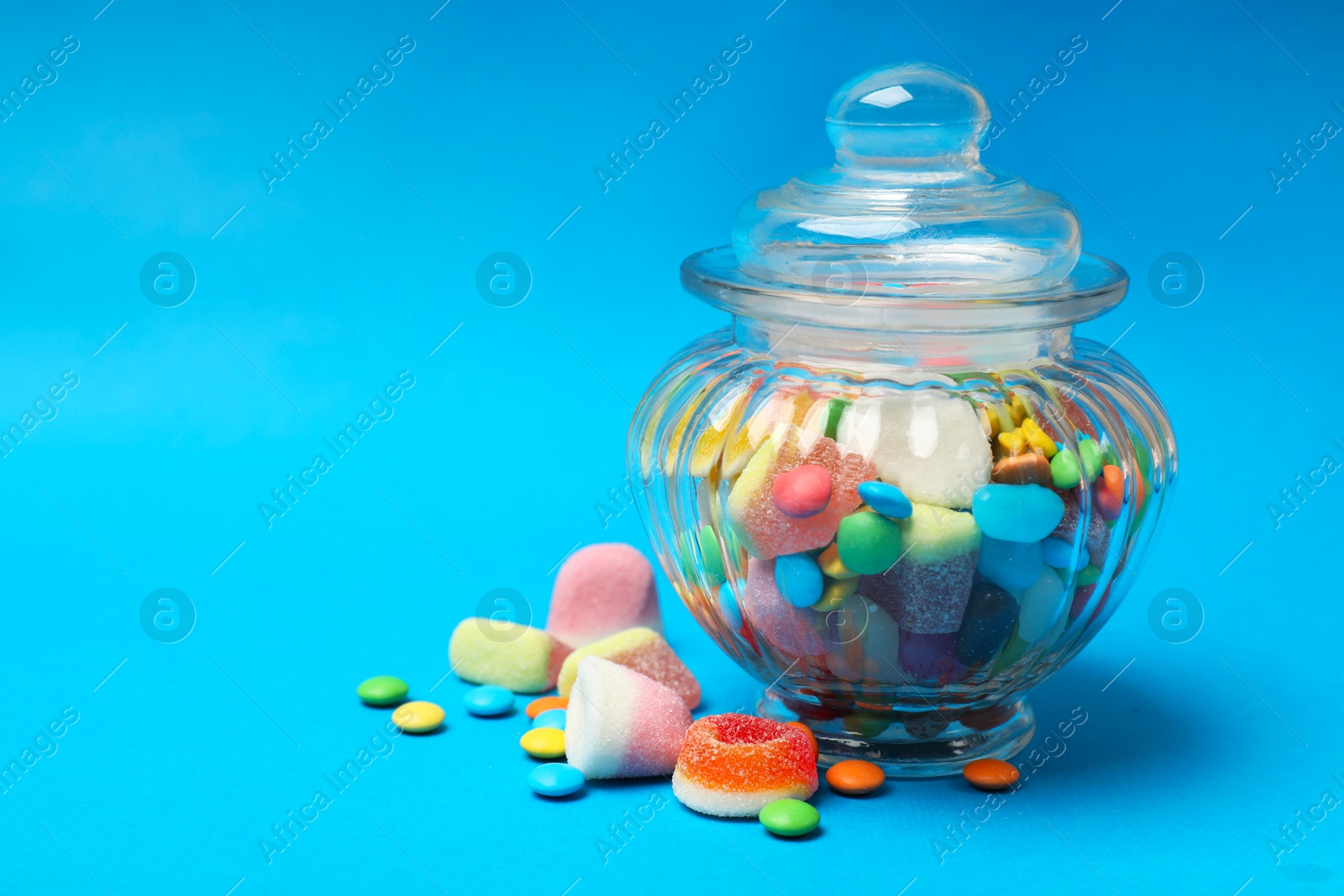 Photo of Different tasty candies in glass jar on light blue background, closeup. Space for text