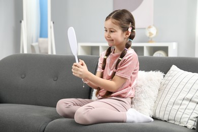 Photo of Little girl with beautiful hair accessories looking into mirror on sofa at home