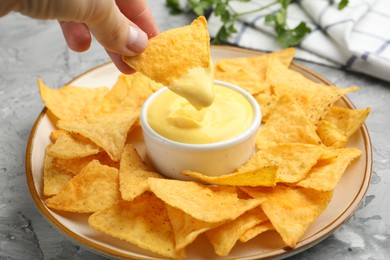 Photo of Woman dipping nacho chip into tasty cheese sauce at grey table, closeup
