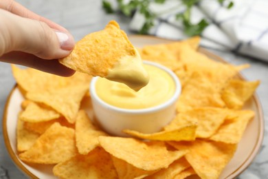 Photo of Woman dipping nacho chip into tasty cheese sauce at table, closeup