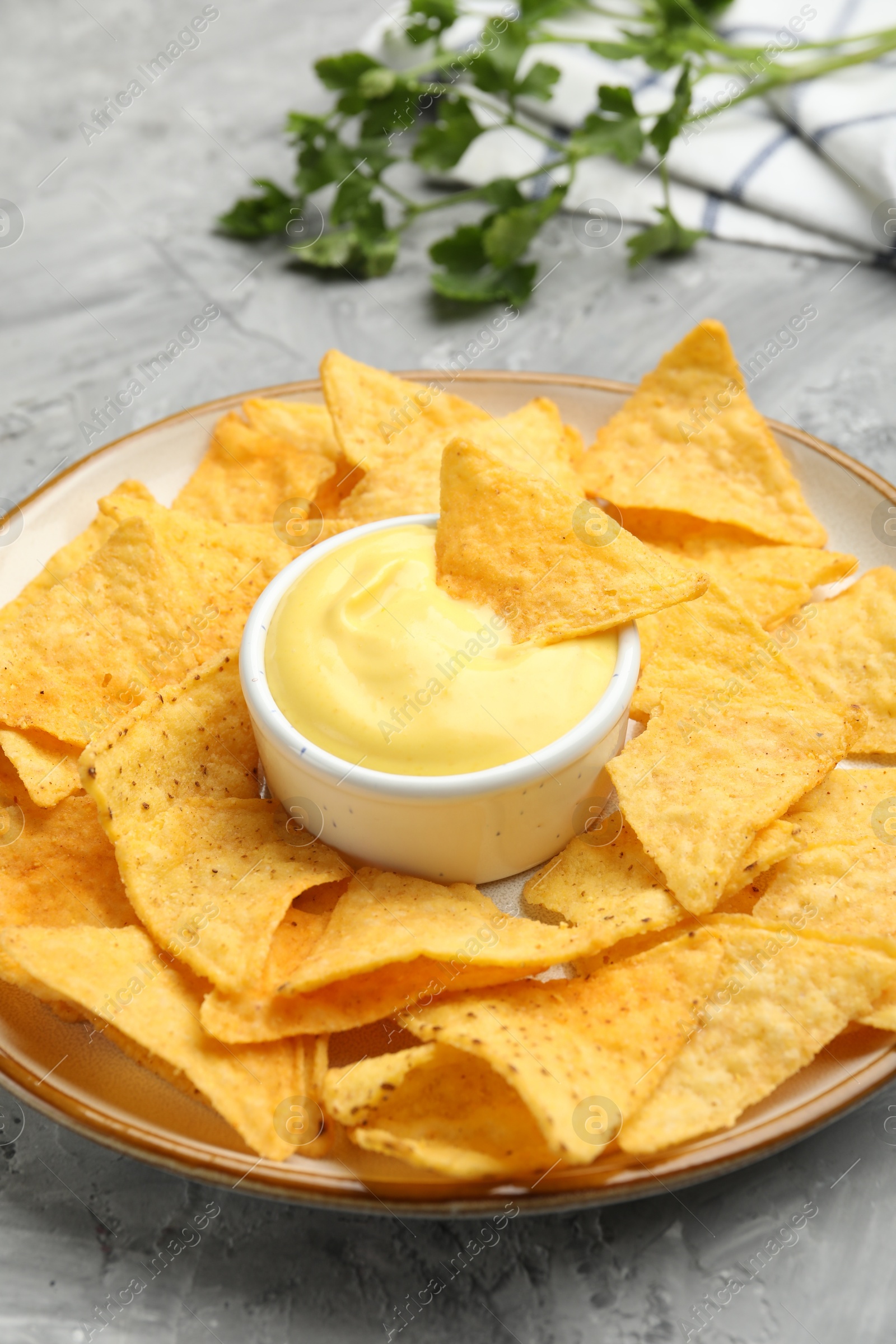 Photo of Tasty cheese dipping sauce in bowl, nacho chips and parsley on grey table, closeup