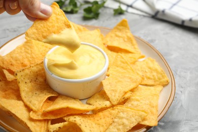 Photo of Woman dipping nacho chip into tasty cheese sauce at grey table, closeup
