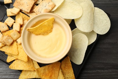 Photo of Tasty cheese dipping sauce in bowl and different snacks on dark wooden table, top view