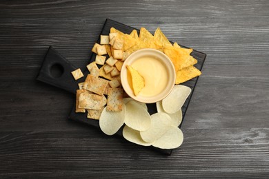 Photo of Tasty cheese dipping sauce in bowl and different snacks on dark wooden table, top view