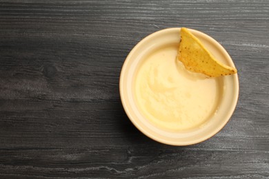 Photo of Tasty cheese dipping sauce in bowl and nacho chip on dark wooden table, top view. Space for text