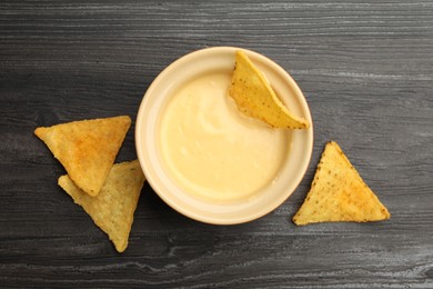 Photo of Tasty cheese dipping sauce in bowl and nacho chips on dark wooden table, flat lay