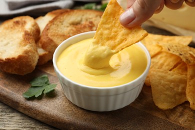 Photo of Woman dipping nacho chip into tasty cheese sauce at table, closeup