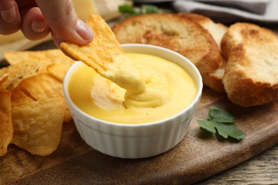 Photo of Woman dipping nacho chip into tasty cheese sauce at table, closeup