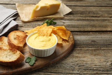 Photo of Tasty cheese dipping sauce in bowl, nacho chips and croutons on wooden table, closeup. Space for text