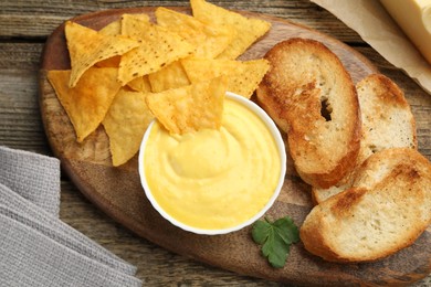 Photo of Tasty cheese dipping sauce in bowl, nacho chips and croutons on wooden table, top view