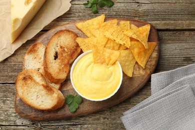 Photo of Tasty cheese dipping sauce in bowl, nacho chips and croutons on wooden table, flat lay