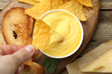 Photo of Woman dipping nacho chip into tasty cheese sauce at wooden table, top view