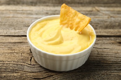 Photo of Tasty dipping cheese sauce in bowl and nacho chip on wooden table, closeup