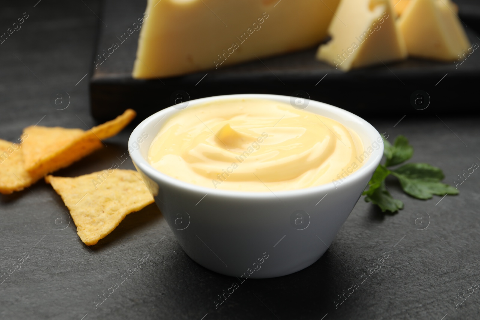 Photo of Tasty dipping cheese sauce in bowl, nacho chips and parsley on black table, closeup