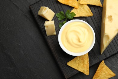 Photo of Tasty dipping cheese sauce in bowl, nacho chips and parsley on black table, top view. Space for text