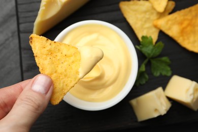 Photo of Woman dipping nacho chip into tasty cheese sauce at table, top view