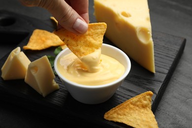 Photo of Woman dipping nacho chip into tasty cheese sauce at black table, closeup
